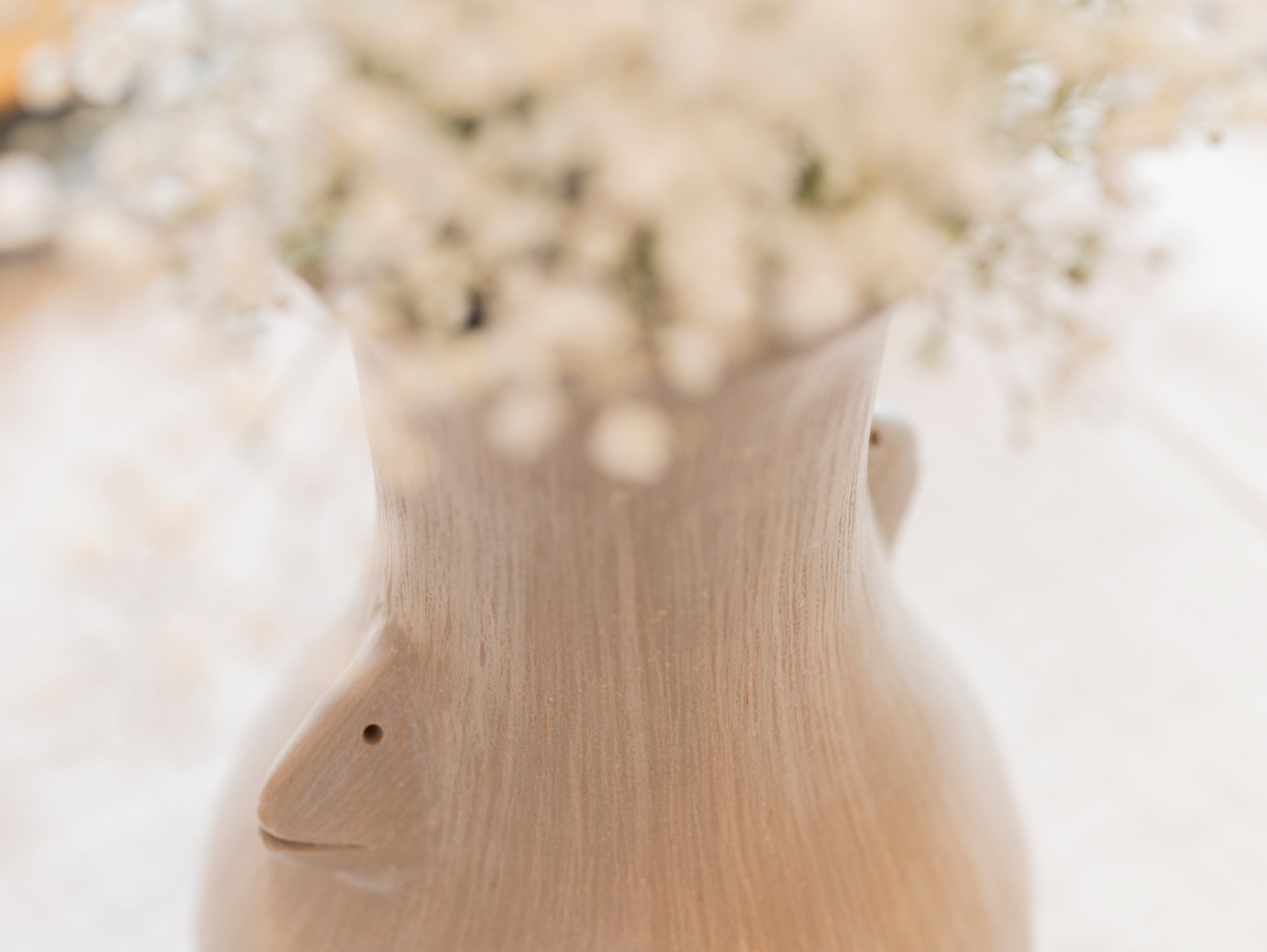 Close-up of a handcrafted ceramic vase with sculpted face detail, holding delicate white baby’s breath flowers on a linen-covered table.